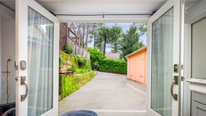 Blick durch eine Glastür in einen überdachten Außenbereich mit einem Betonweg, Grünflächen und einem Holzzaun.