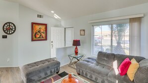 A living room with a sofa, ottoman, and a coffee table. There is a lamp on a side table, a clock on the wall, and a window with curtains. - Victorville Home w/ Fenced Backyard + Patio! (Victorville)
