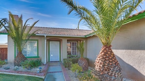 A single-story house with a tiled roof, a front door, and a small front yard with palm trees and shrubs. - Victorville Home w/ Fenced Backyard + Patio! (Victorville)
