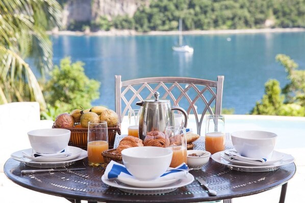 Disposição de mesa de café da manhã com croissants, frutas e bebidas, ao lado de uma piscina com vista panorâmica.
