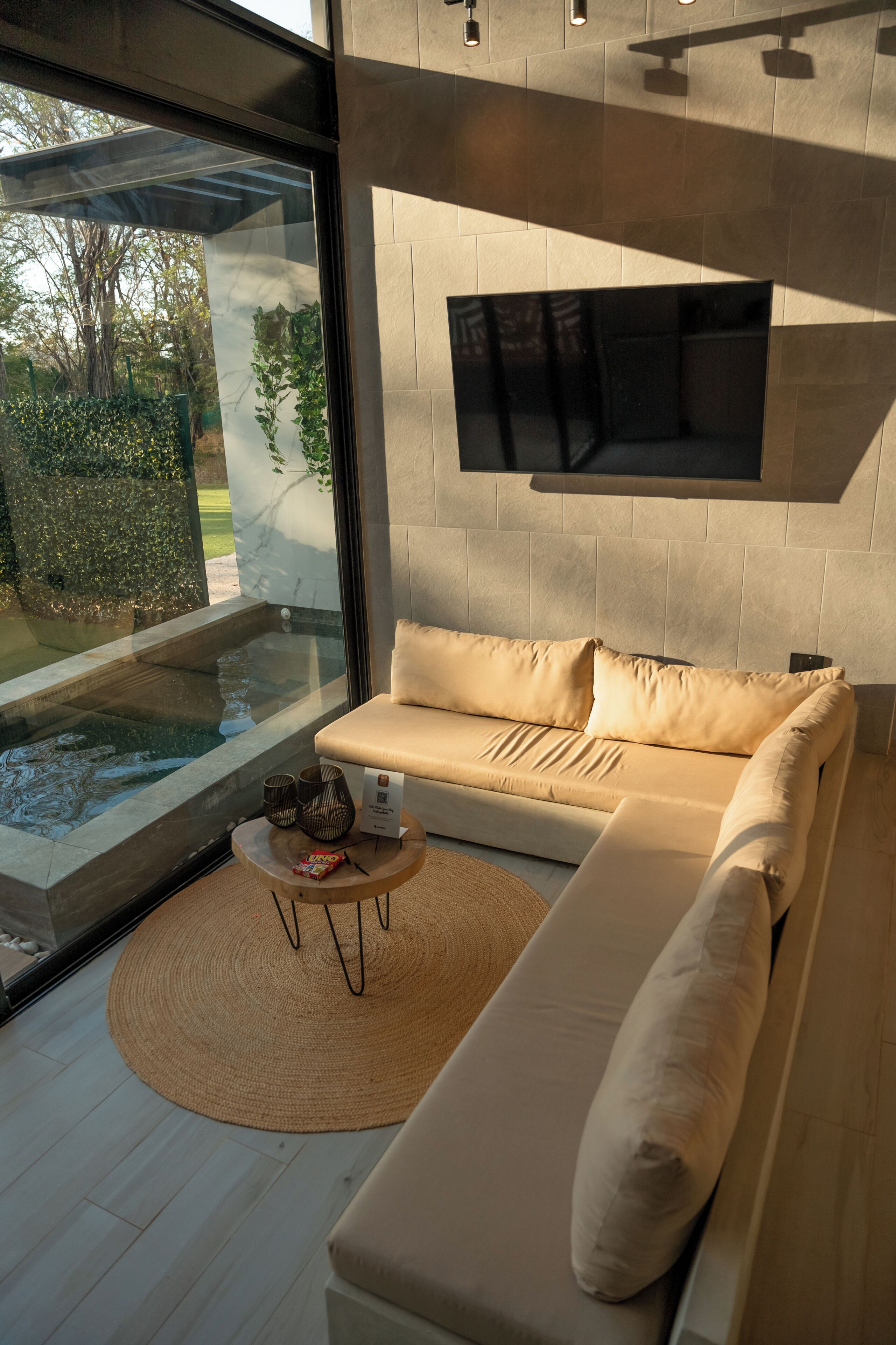 Ein modernes Wohnzimmer mit einem beigen Sofa, einem an der Wand befestigten Flachbildfernseher, einem kleinen runden Tisch und einem gewebten Teppich sowie einer Glaskonstruktion mit Wasserspiel.