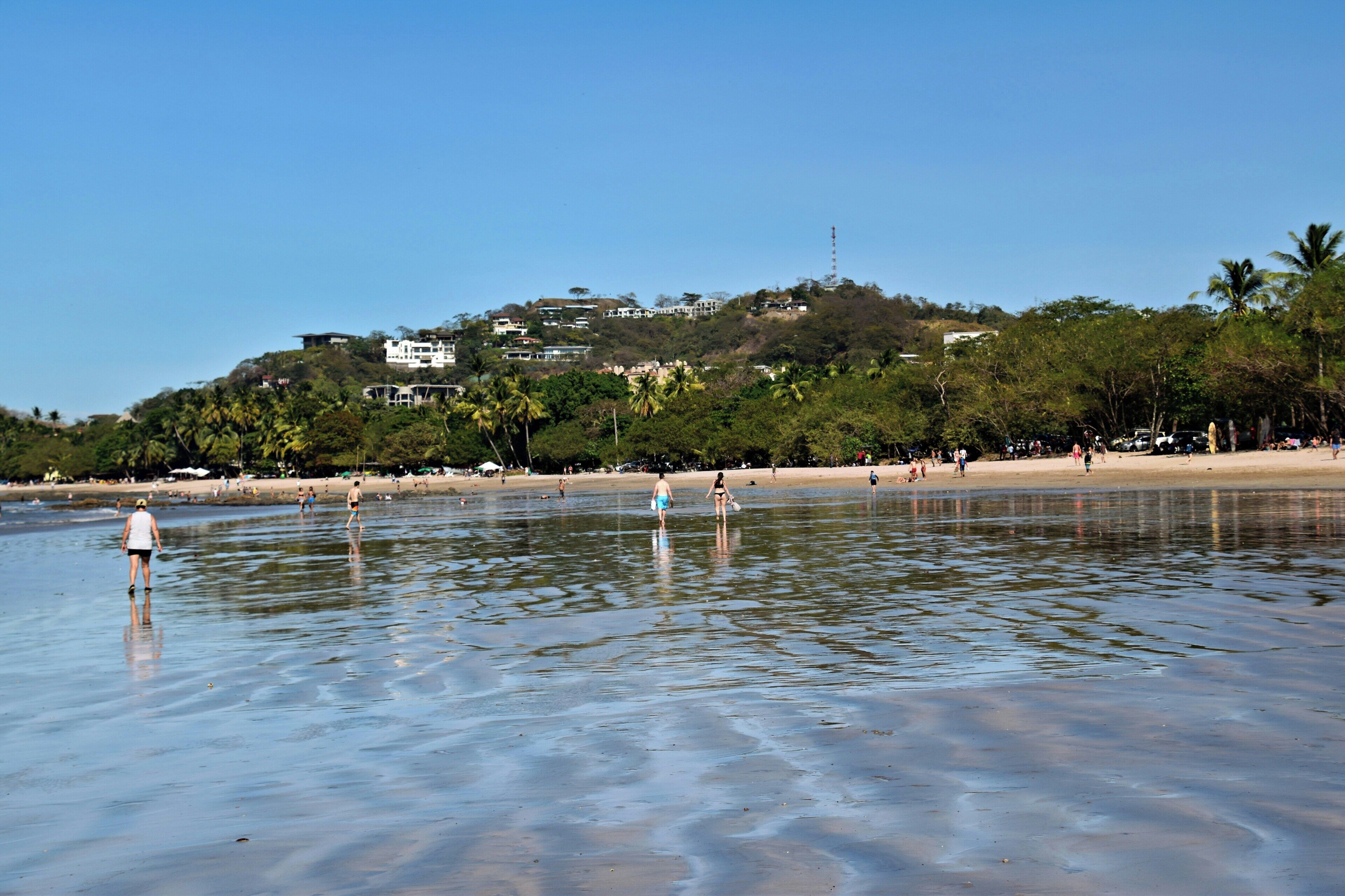 A beach with people walking, a clear sky, and buildings on a hill in the background.