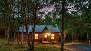 Metallikatolla varustettu hirsihökkelö, jonka ympärillä on puita, ja jonka edessä on kyltti, josta ilmenee, että kyseessä on mökki.