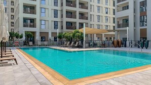 A swimming pool with lounge chairs and umbrellas, surrounded by a multi-story building with balconies. - Creek Beach Roomy 3BR with terrace (Dubai)