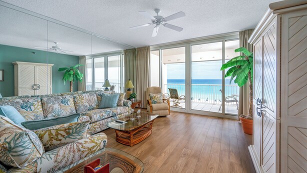 A spacious living room with a large sofa, a coffee table, and a chair. There is a ceiling fan, a large window with a view of the ocean, and a potted plant. - Relax in 409...Get in a Flip-Flop State of Mind (Navarre)