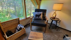 A room with a view of trees through a window, a chair with a cow pillow, a lamp on a side table, a wicker basket with clothes, and a small wooden table. - Adventure Cabin -Coolin Priest Lake; king bed, bunks, ATV and snowmobile parking (Coolin)