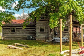 Une cabane rustique en rondins de bois, surmontée d’un toit en métal, entourée d’une cour verdoyante et dotée d’une terrasse en bois avec des tables à carreaux.