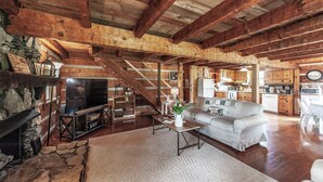 A rustic cabin interior with a stone fireplace, wooden beams, a living room with a sofa and coffee table, a kitchen area, and a staircase leading to an upper level.