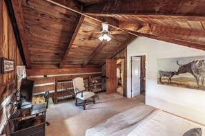 Une pièce rustique avec des poutres en bois, un ventilateur de plafond, un tableau représentant une vache et une chaise rustique.