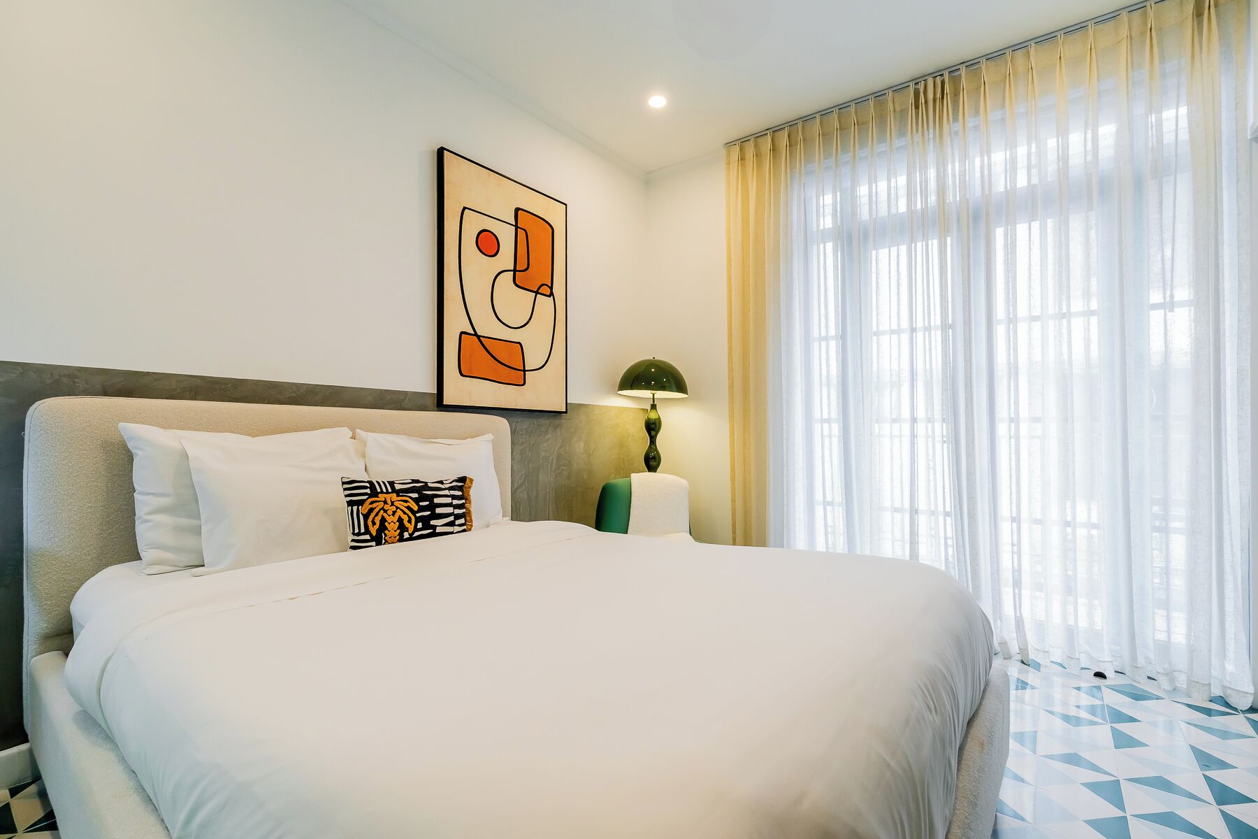 Habitación de hotel con una cama grande, un cuadro en la pared, una lámpara y una ventana con cortinas.