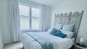 Un dormitorio con una cama grande, una ventana con cortinas, una mesita de noche con lámpara y un cabecero decorativo.
