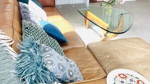 Una sala moderna con un sofá de cuero marrón, una mesa de centro de vidrio y un comedor con una isla blanca y taburetes de bar.
