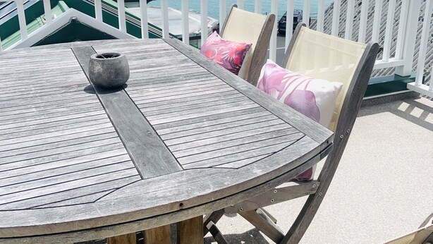 Una mesa de madera con sillas y una barandilla blanca, con vista a un mar tranquilo y barcos.