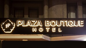 The exterior of Plaza Boutique Hotel with a neon sign, a red carpet, and a glass entrance.