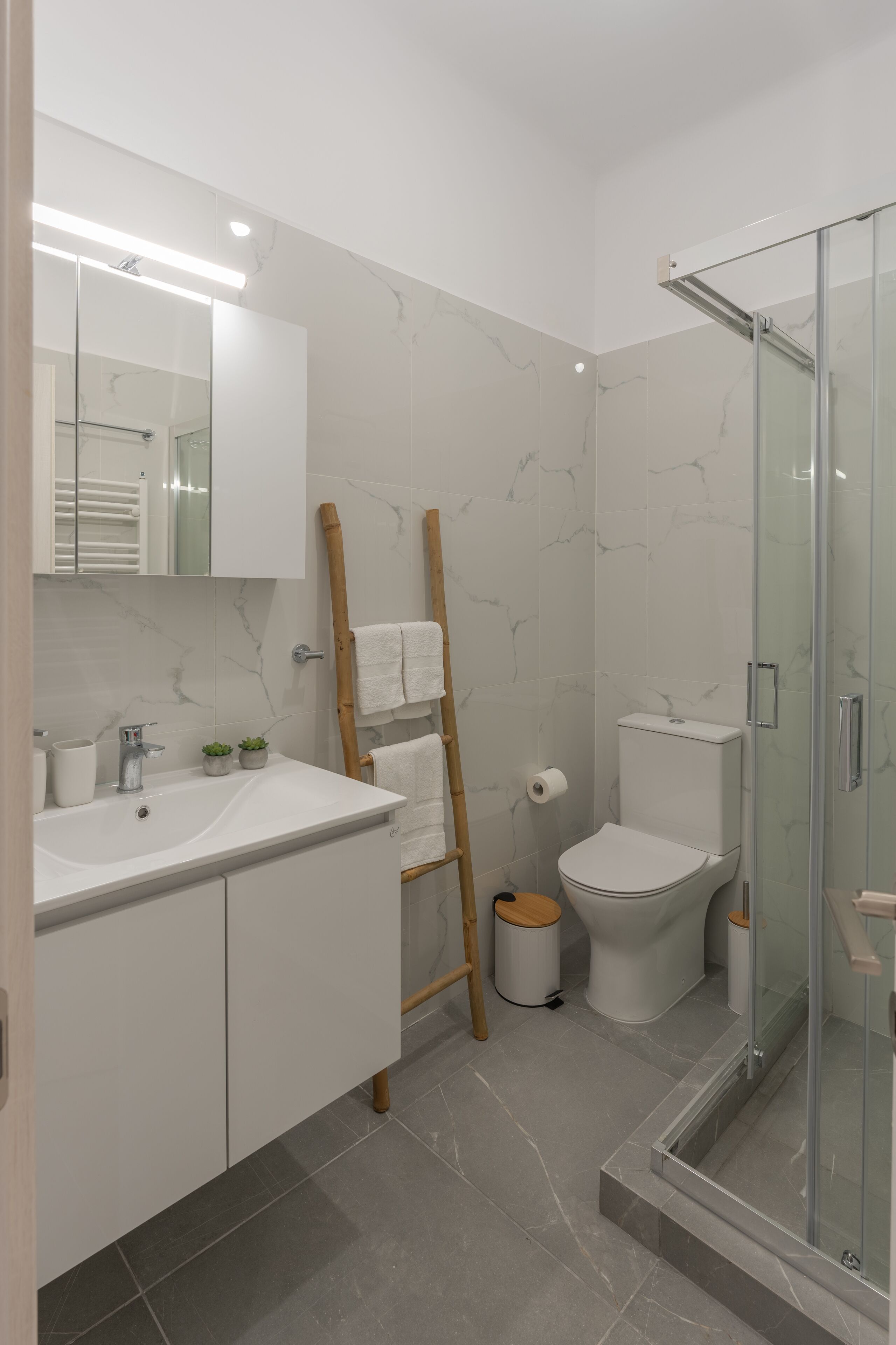 Een moderne badkamer met een glazen douchecabine, een wit wastafelmeubel met spiegel erboven en een toilet met een marmeren achterwand.