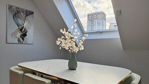 Un comedor moderno con una mesa redonda, sillas color beige, un jarrón con flores secas, un cuadro abstracto enmarcado y vistas de un edificio a través de un tragaluz.