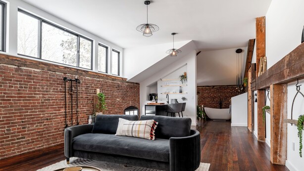 A modern living room with a dark sofa, a white rug, and a brick wall. There is a large window, a hanging light fixture, and a wooden beam structure on the right. - Abacus Suite at Spofford Lodge (Bucksport)