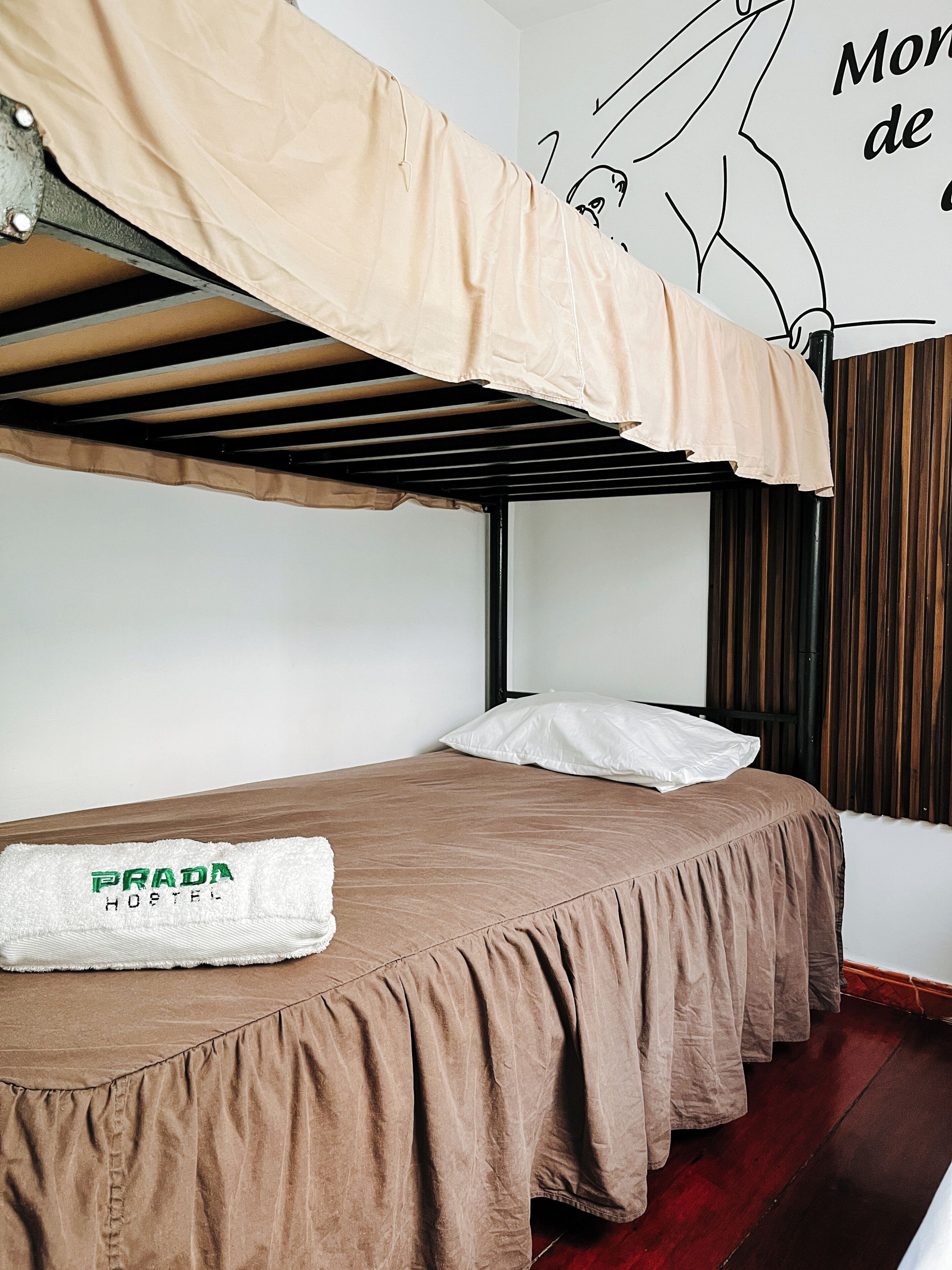 Een stapelbed met een bruine sprei en een wit kussen, een handdoek met de tekst 'PRADA HOSTEL' en een muurschildering.