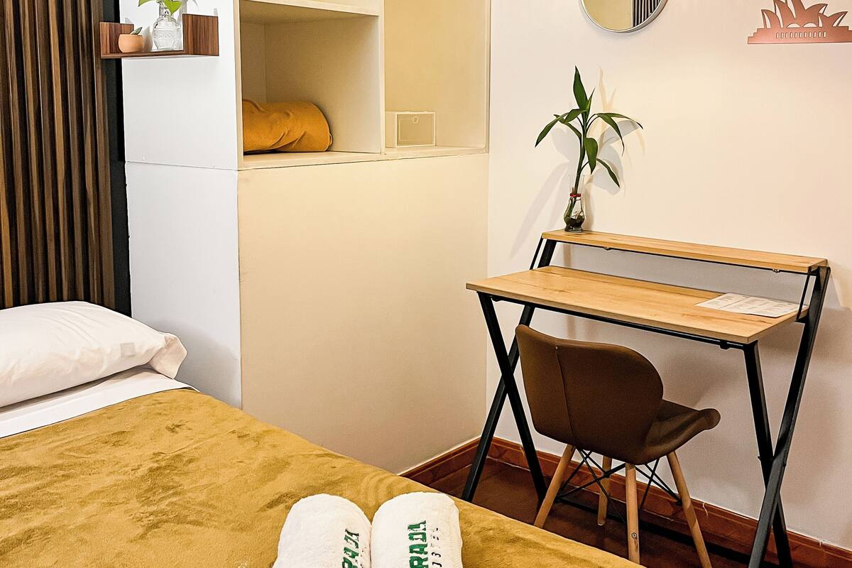 Une chambre d’hôtel comprenant un lit, un bureau, une chaise, une armoire, un luminaire suspendu, un miroir et une plante.
