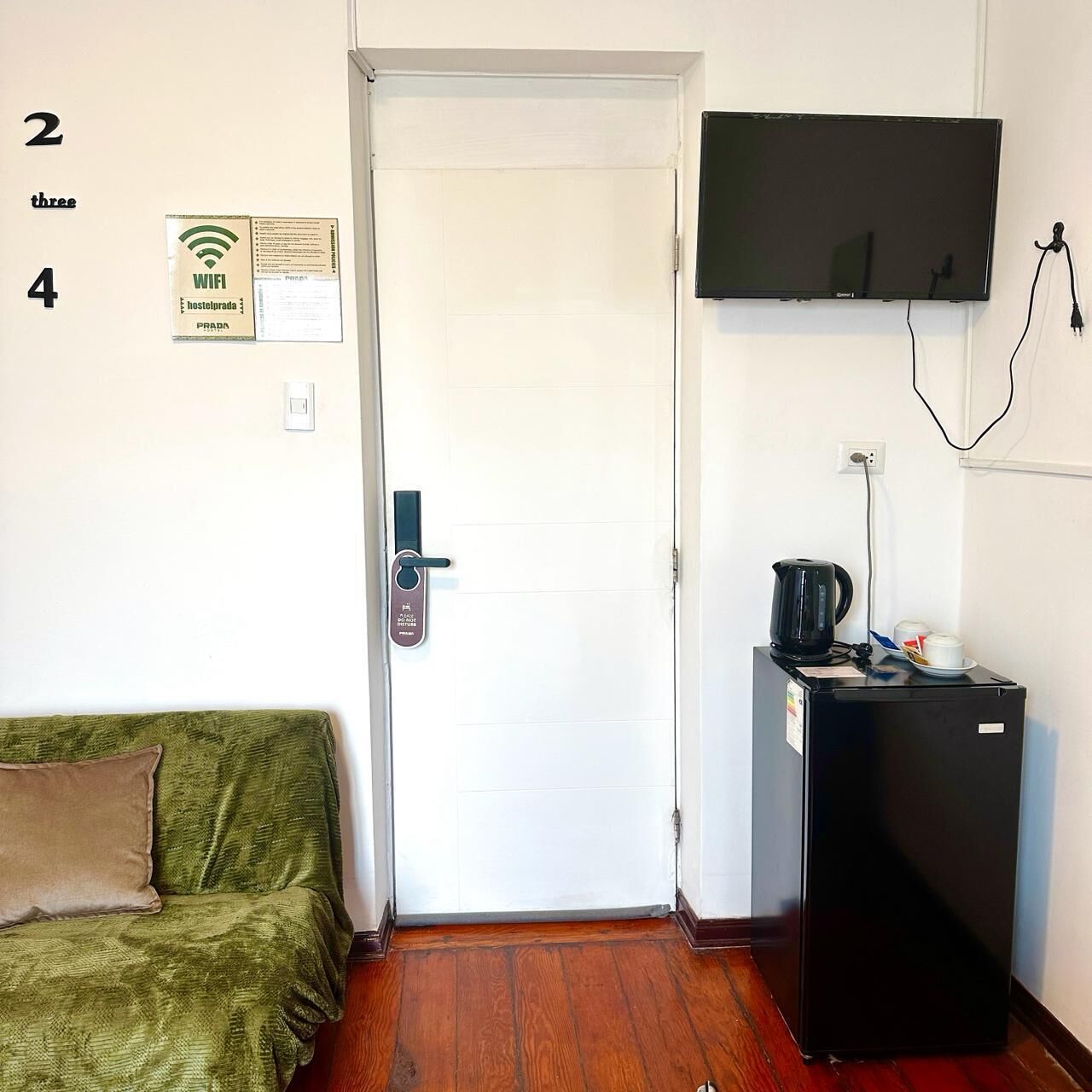 A room with a white door, a green sofa, a black refrigerator, a television mounted on the wall, and a WiFi sign on the wall.