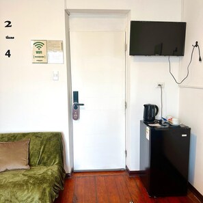 A room with a white door, a green sofa, a black refrigerator, a television mounted on the wall, and a WiFi sign on the wall.