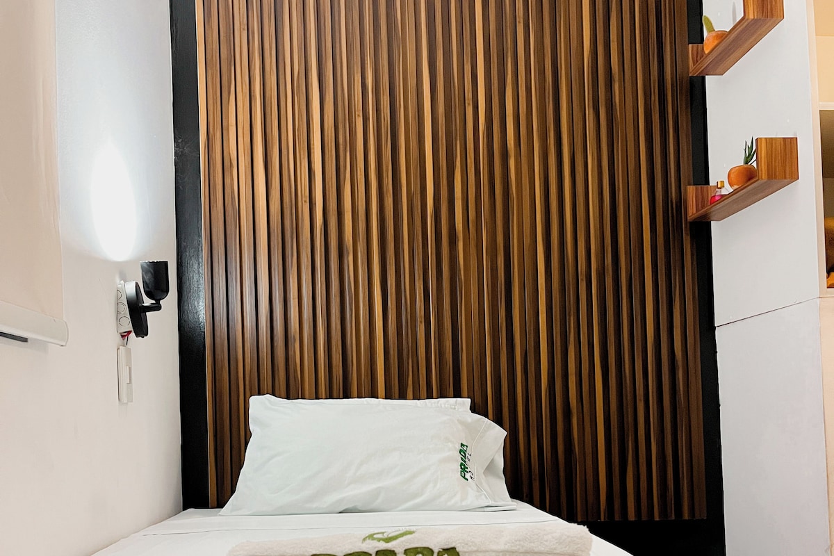 Una cama con una colcha blanca y una toalla con la marca 'PRADA HOTEL', una pared de listones de madera y un pequeño estante de madera con una planta.