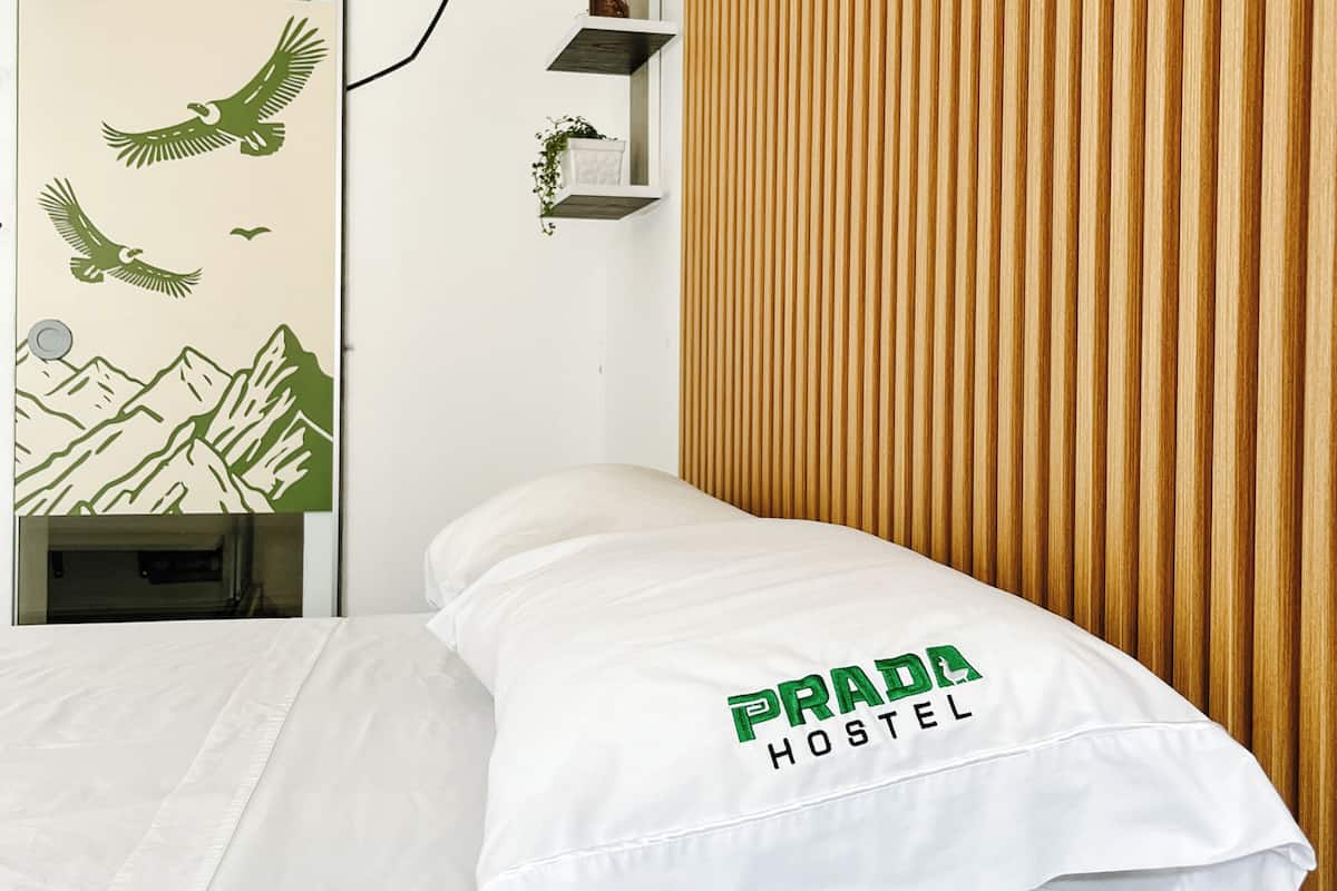 Un lit avec un oreiller portant l’inscription « PRADA HOSTEL », un mur avec des lambris en bois verticaux et une plante suspendue.