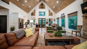 A spacious living area with a high ceiling, wooden beams, a brown leather sofa, a coffee table, and large windows offering views of the outdoors.