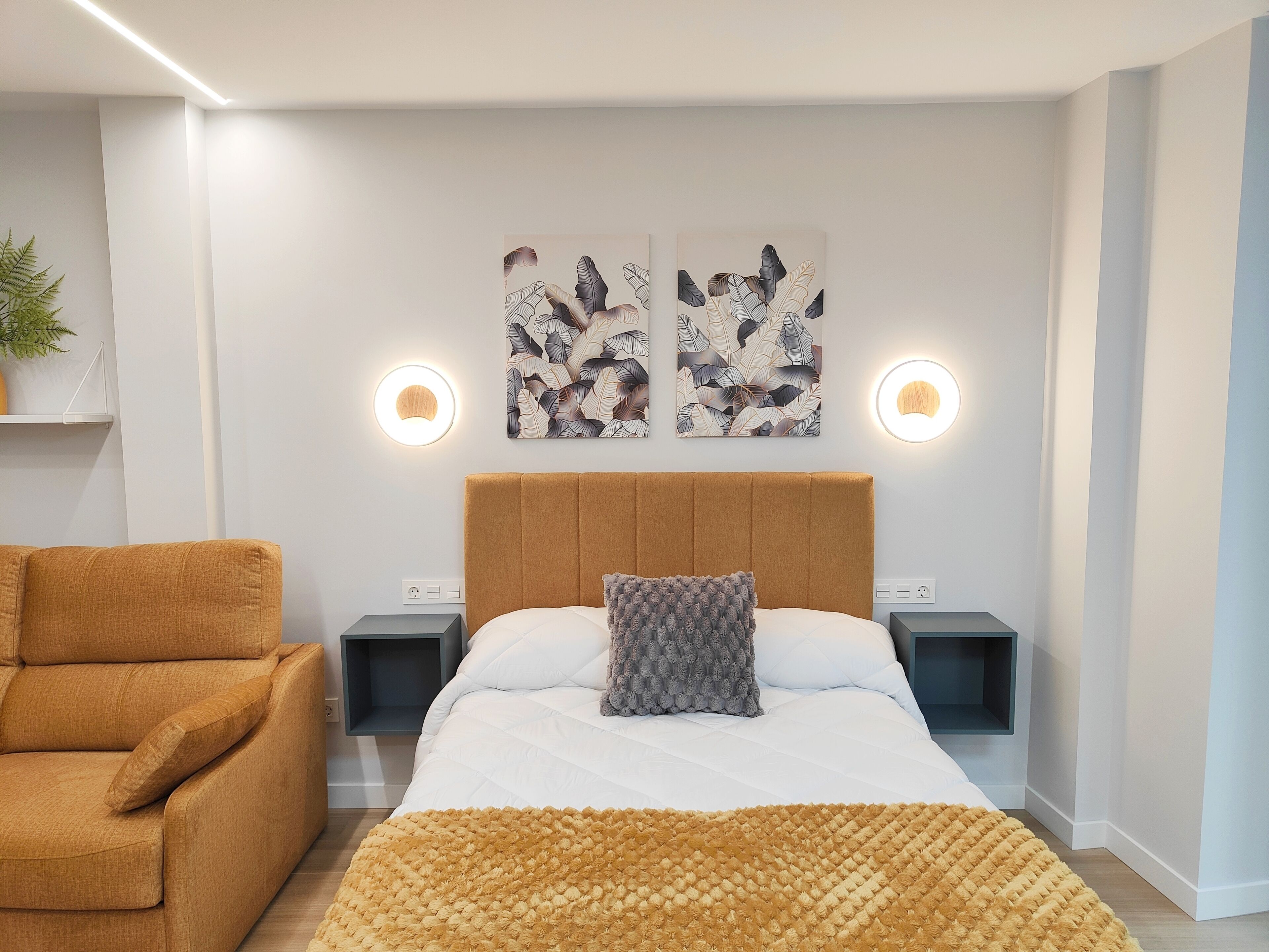 Une chambre moderne avec un grand lit, un fauteuil marron, deux appliques murales et des œuvres d’art ornées de motifs de feuilles.