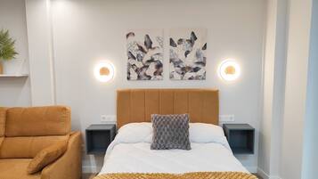 Une chambre moderne avec un grand lit, un fauteuil marron, deux appliques murales et des œuvres d’art ornées de motifs de feuilles.