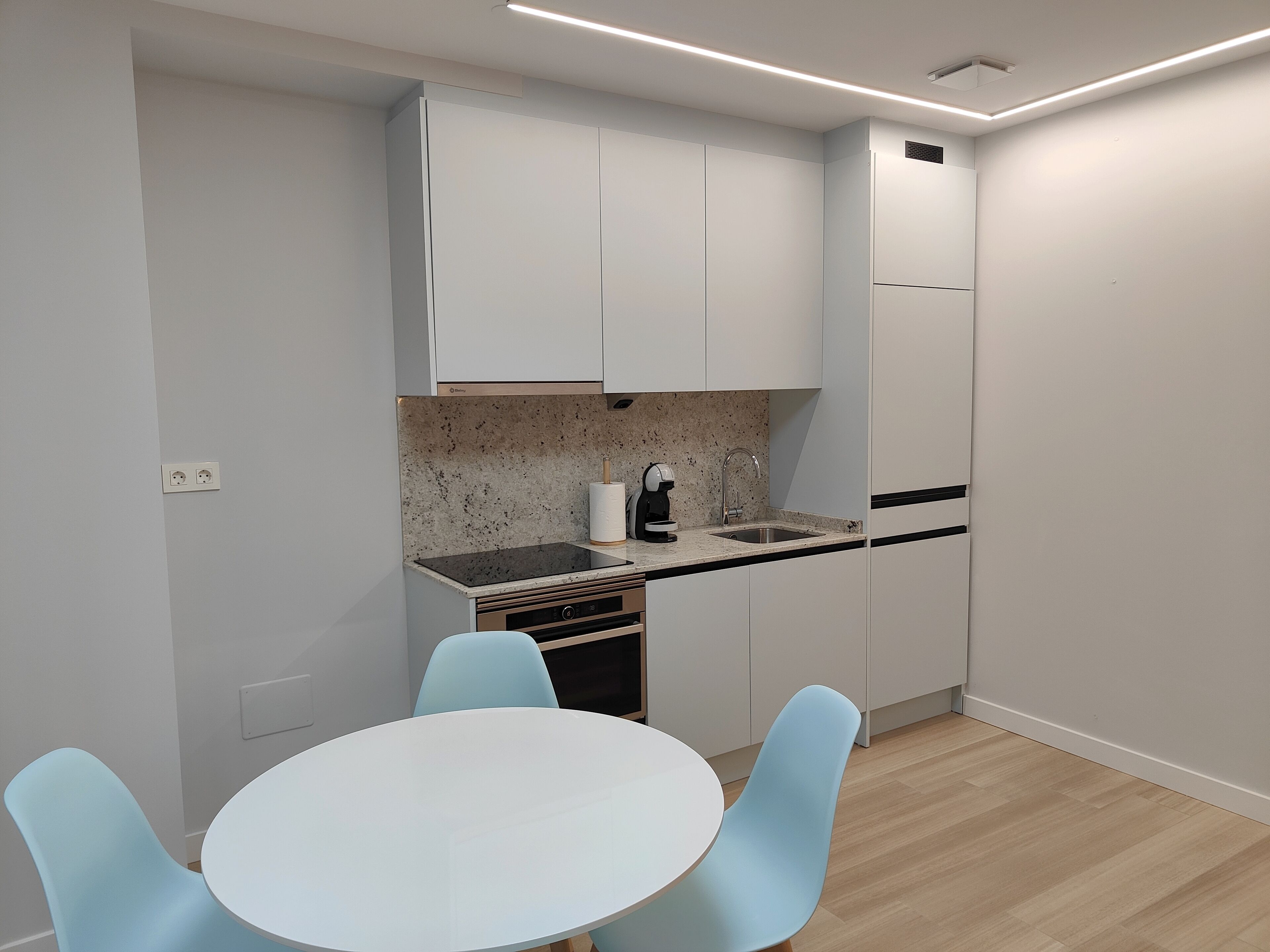 Une cuisine moderne avec des armoires blanches, un comptoir en granit, un four encastré, un évier et une table ronde avec des chaises bleues.