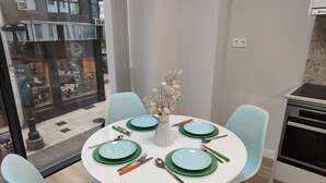 Et spiseområde med et rundt bord dækket til fire personer, lyseblå stole og en vase med tørrede blomster.