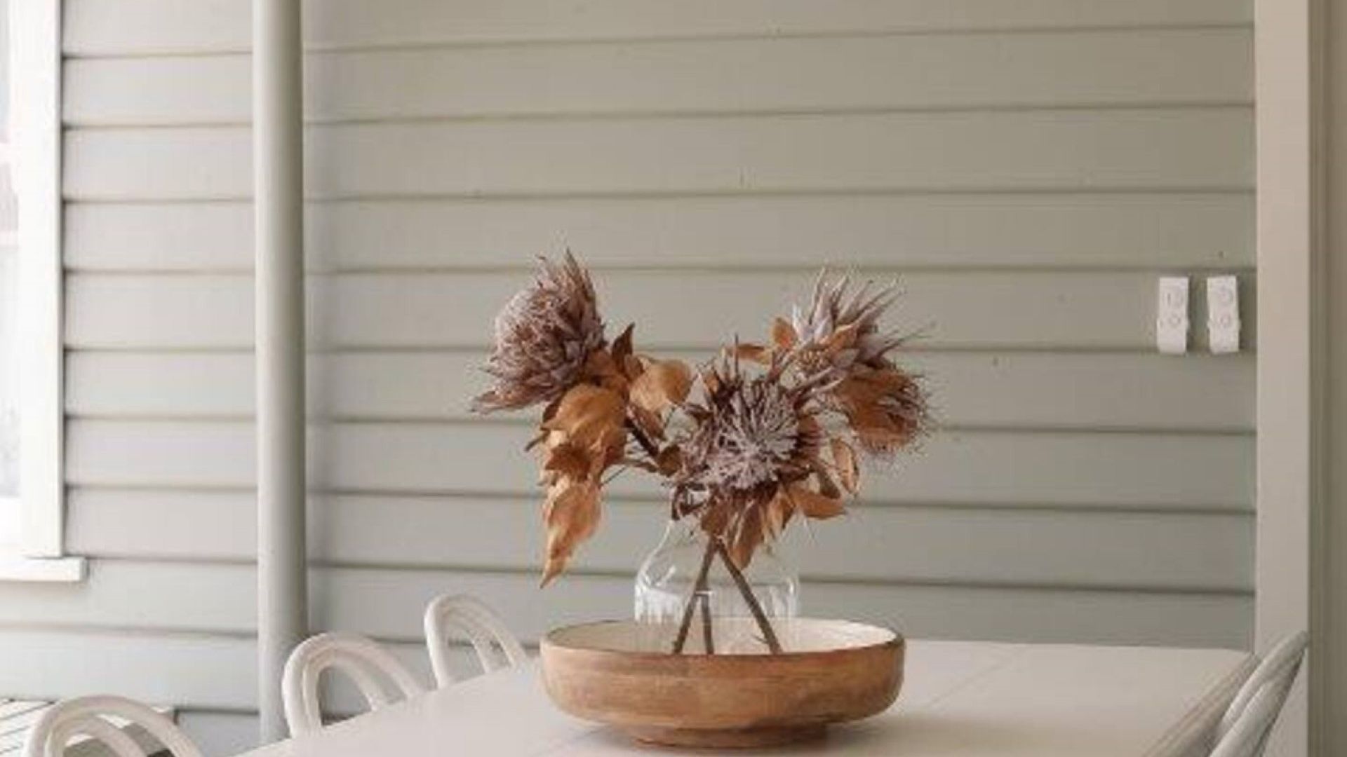 Una mesa con un cuenco de madera con flores secas, frente a una pared blanca con revestimiento horizontal.