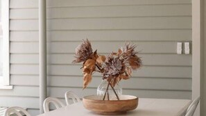 A table with a wooden bowl containing dried flowers, set against a white wall with horizontal siding.