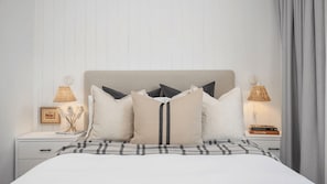 Una cama bien hecha con colcha escocesa, cabecera y varios almohadones. Hay dos mesitas de noche con lámparas y un cuadro enmarcado en una de ellas.
