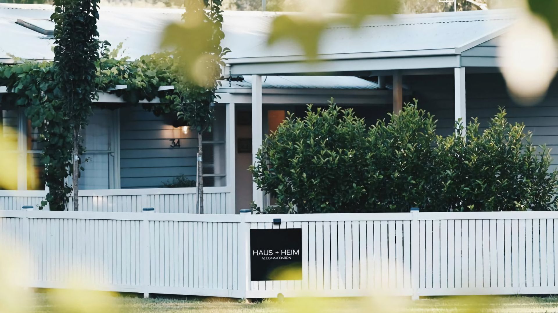 A building with a white fence, a sign reading 'HAUS + HEIM ACCOMMODATION', and a covered porch area.