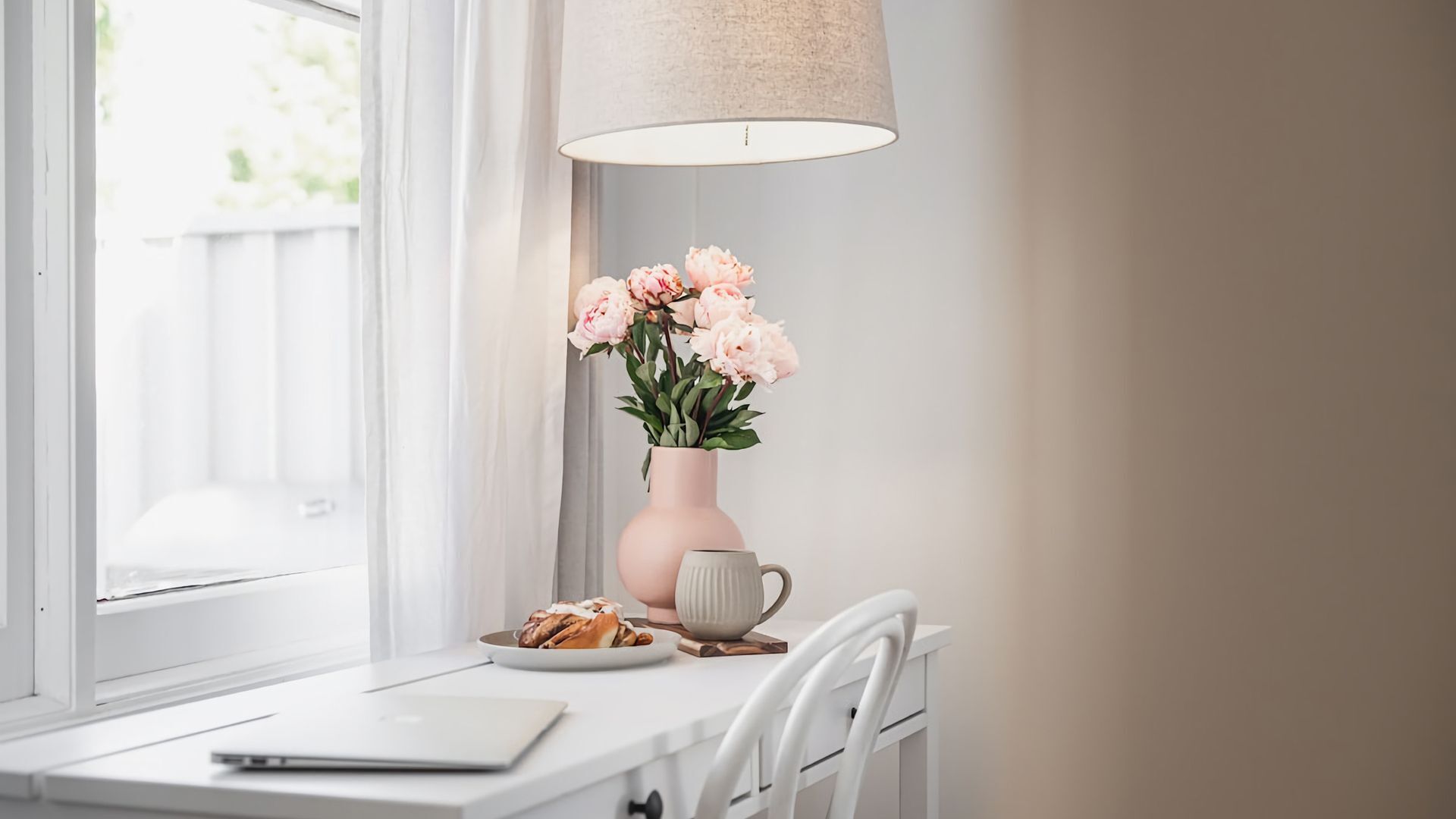 A white desk with a laptop, a plate of pastries, a vase with flowers, and a lamp.