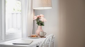 Un escritorio blanco con una computadora portátil, un plato de pasteles, un florero con flores y una lámpara.