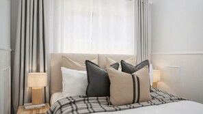 A neatly made bed with a plaid bedspread and multiple pillows, a bedside lamp, and a window with sheer curtains.