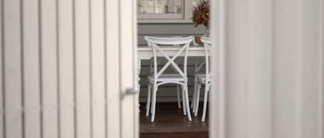 A view through a doorway into a room with a white table and chairs, a window with curtains, and a potted plant.