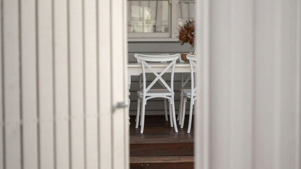 Vista a través de un vano de puerta que da a una habitación con una mesa y sillas blancas, una ventana con cortinas y una planta en maceta.