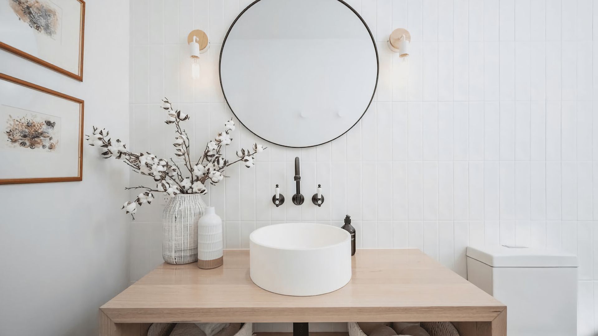 A bathroom with a round mirror, white tiles, a wooden vanity, and two framed artworks.