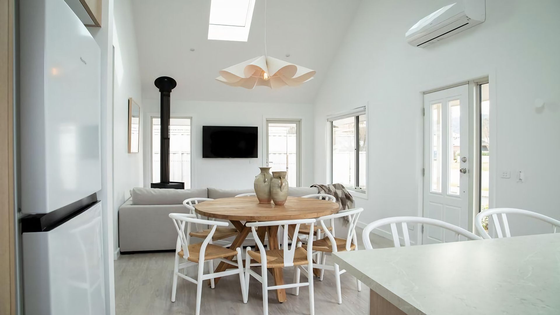 A modern kitchen with white cabinetry, a central island with seating, a flat-screen TV, and a large window providing natural light.