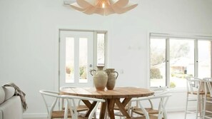 Un espace repas avec une table ronde en bois et des chaises blanches, un luminaire au plafond et une vue dégagée sur l’extérieur grâce à de grandes fenêtres.