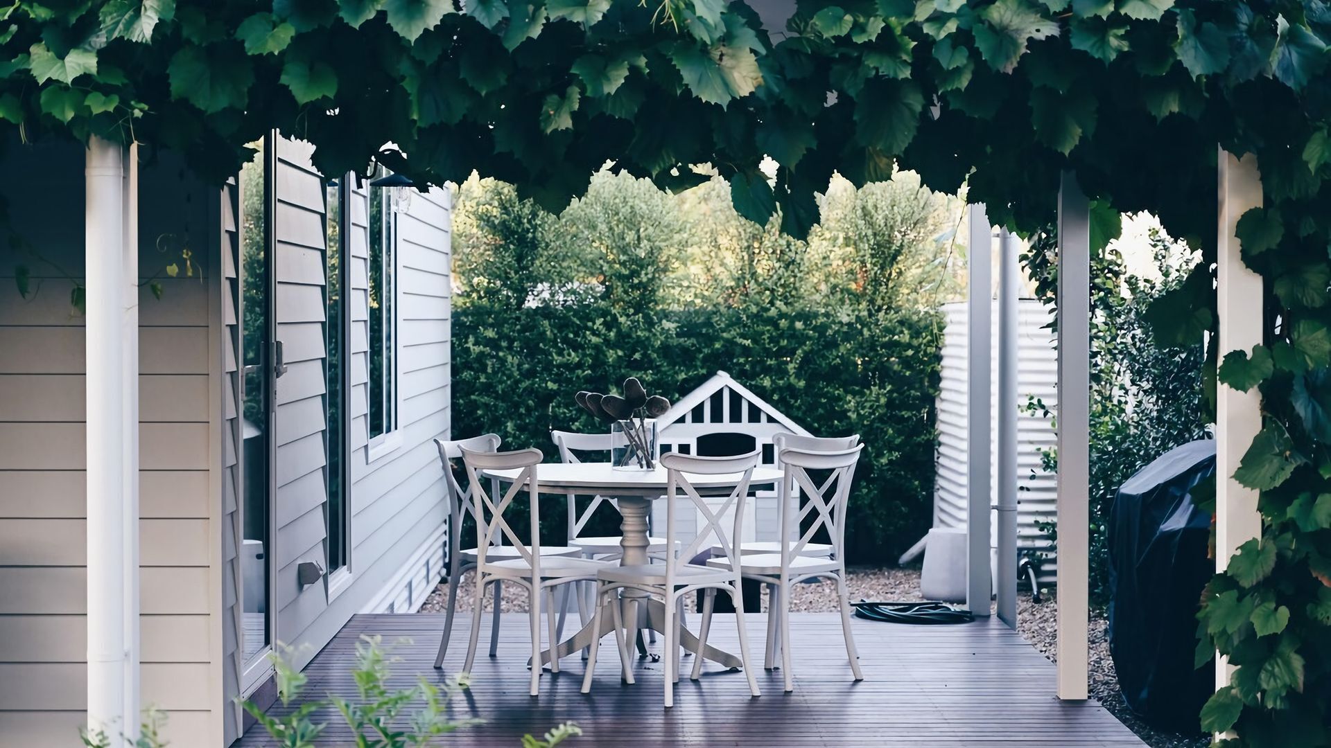 A covered patio with a white table and chairs, surrounded by greenery and a white fence.