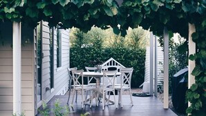 A covered patio with a white table and chairs, surrounded by greenery and a white fence.