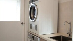 Een compacte wasmachine en droger staan in een kleine wasruimte, met een wasbak en een raam.