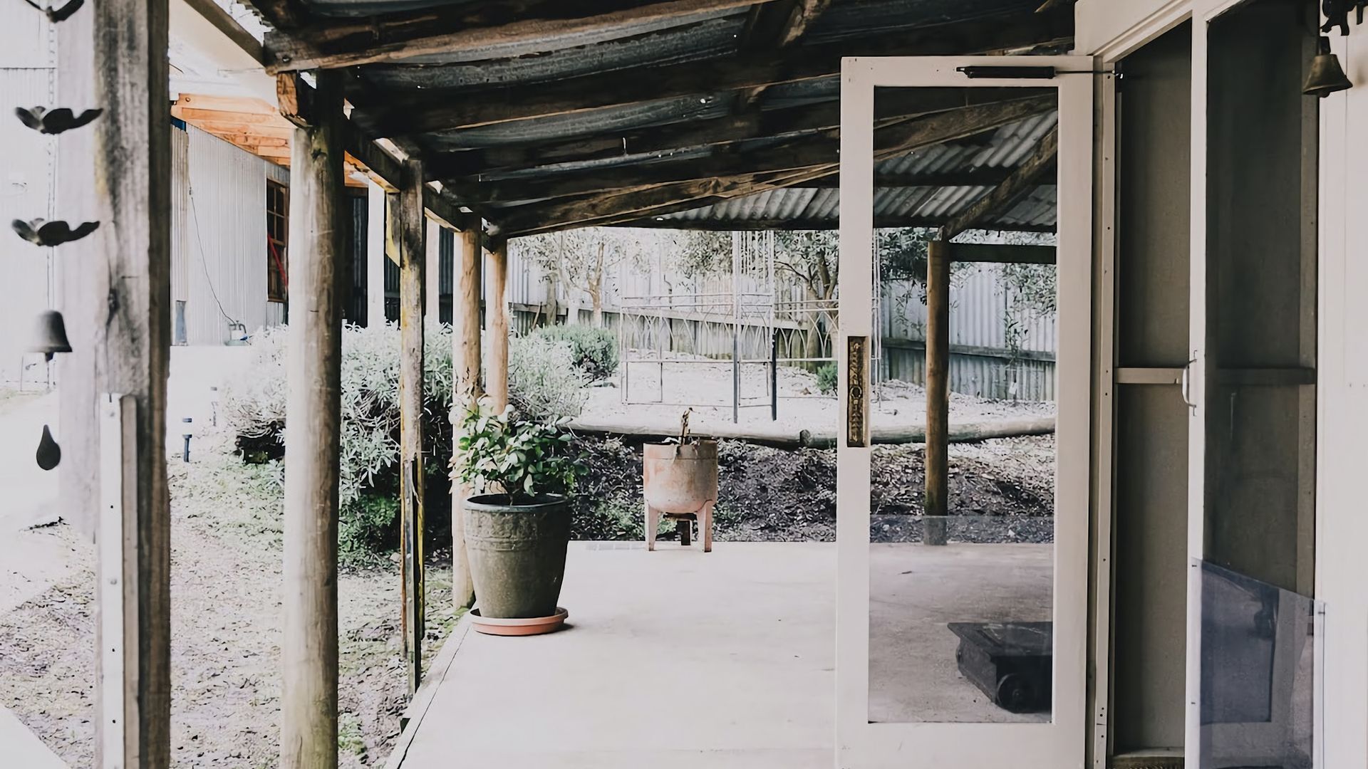 A covered outdoor area with a concrete floor, a large potted plant, and a metal fence in the background.