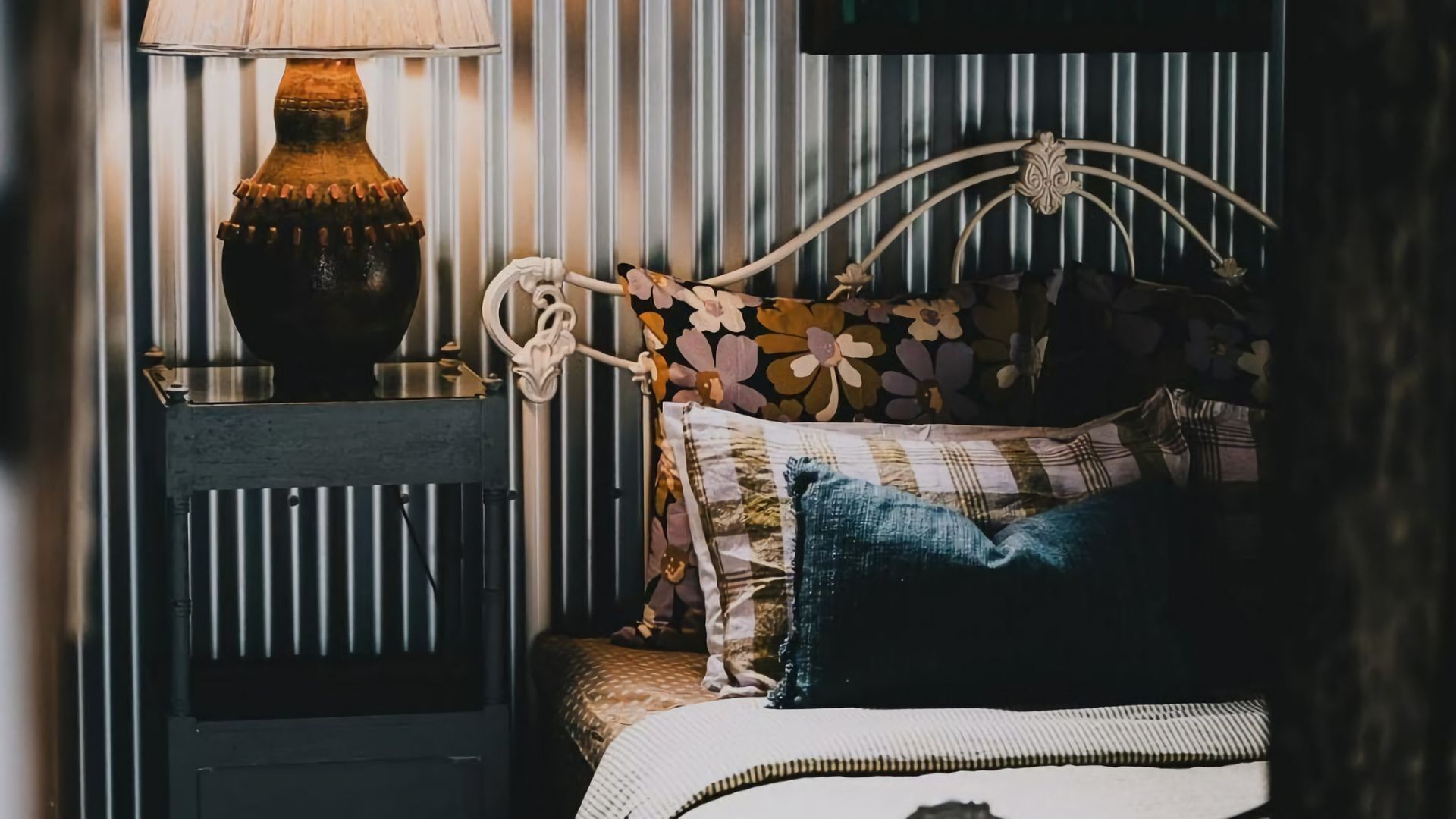 A bed with floral-patterned pillows and a headboard, a bedside table with a lamp, and a wall with vertical stripes.