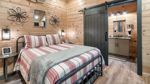 A wooden cabin room with a bed, a small table, and a bathroom visible through a sliding door. - Relaxing Getaway~ K/Q Suites w/ Hot Tub & Fire Pit, Privacy, Great Location (Sevierville)